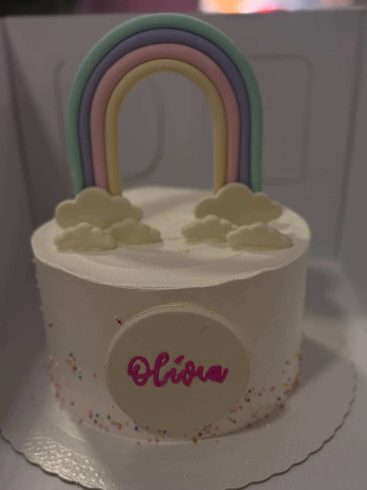 Bolo branco com arco-íris em tons pastel e nuvens no topo, com placa de nome